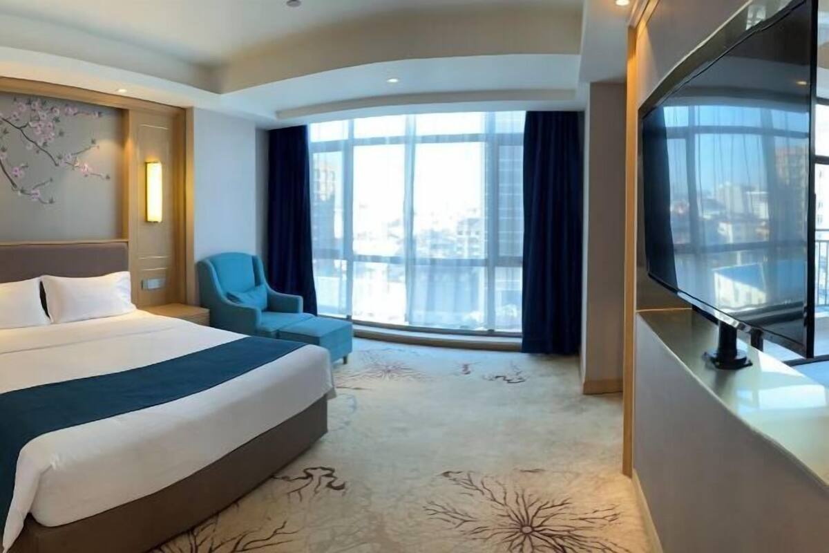 A hotel room with a large bed, a TV, a chair, and a large window with curtains.