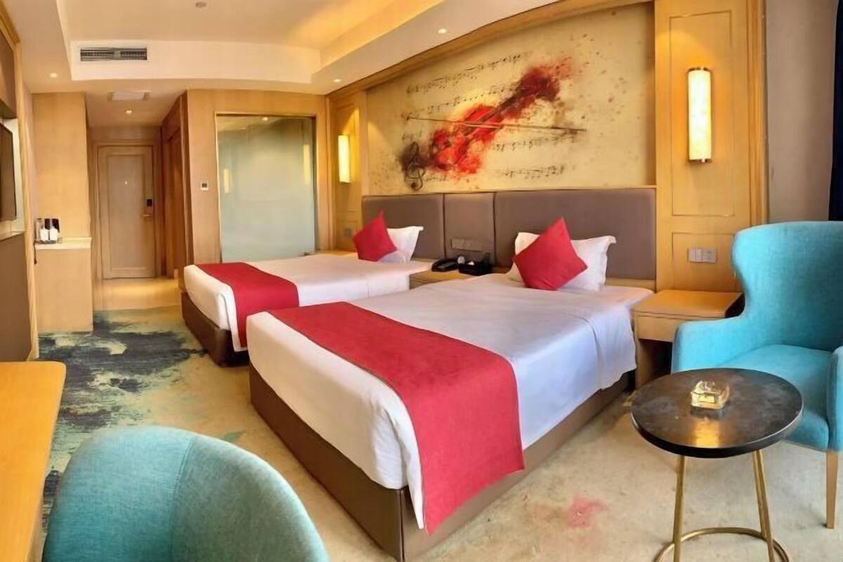 A hotel room with two beds, a small round table, a blue armchair, and a wall with a red abstract painting.
