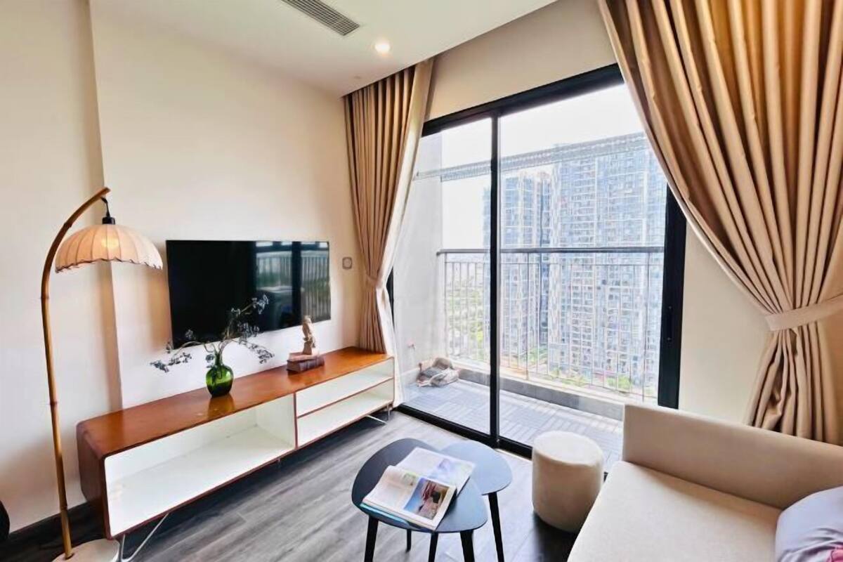 A modern hotel room with a large window, a flat-screen TV, a sofa, a small table, and a lamp.