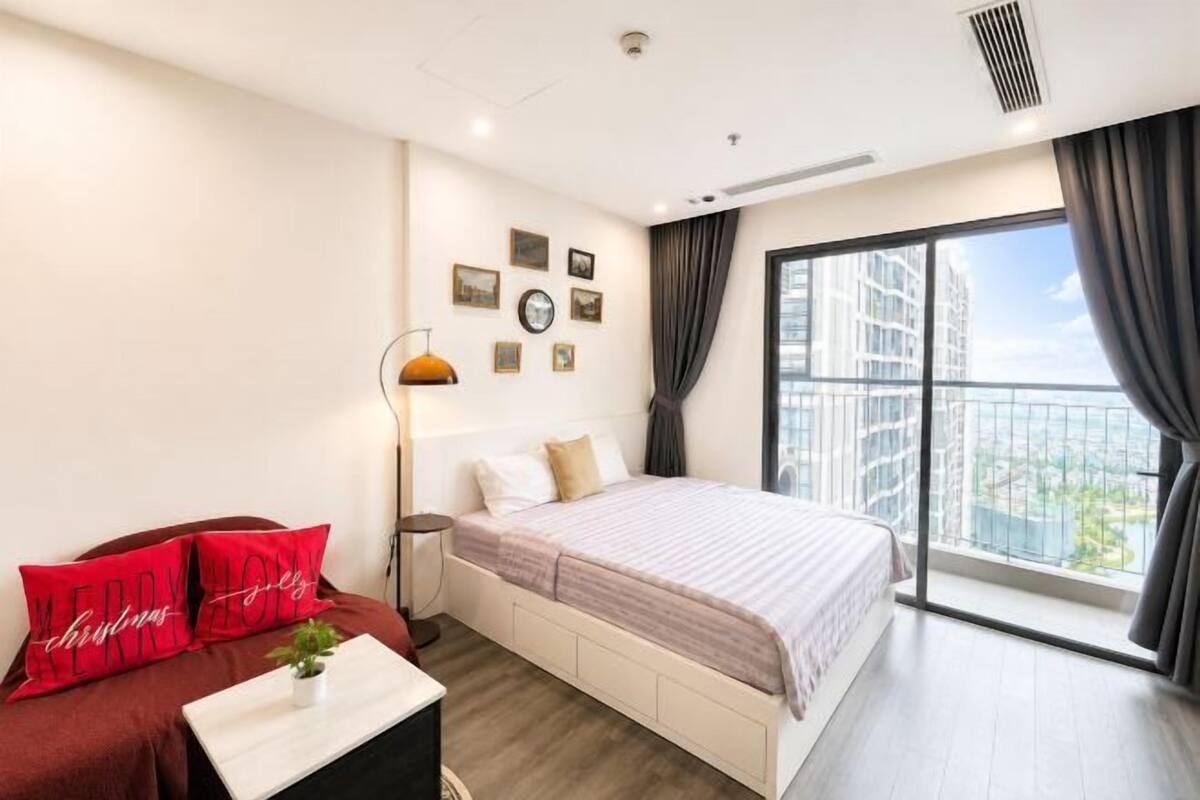 A bedroom with a bed, a bedside table, a lamp, a window with curtains, and a view of buildings.