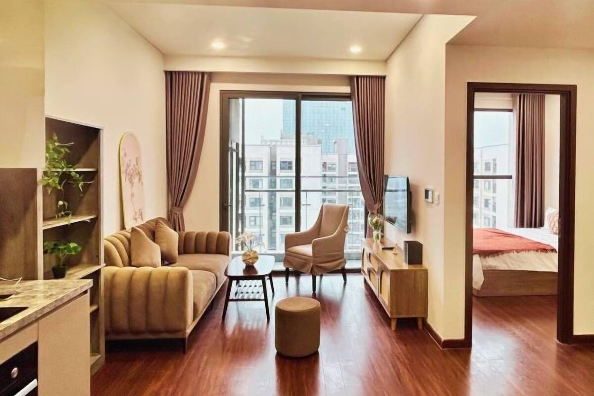 A modern hotel room with a sofa, armchair, ottoman, TV, and a view of the city through large windows.