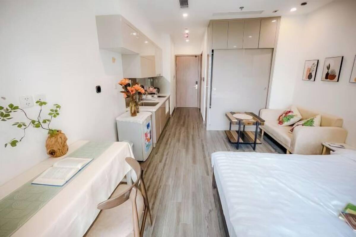 A modern hotel room with a bed, a desk, a chair, a small table, a sofa, and a kitchenette.