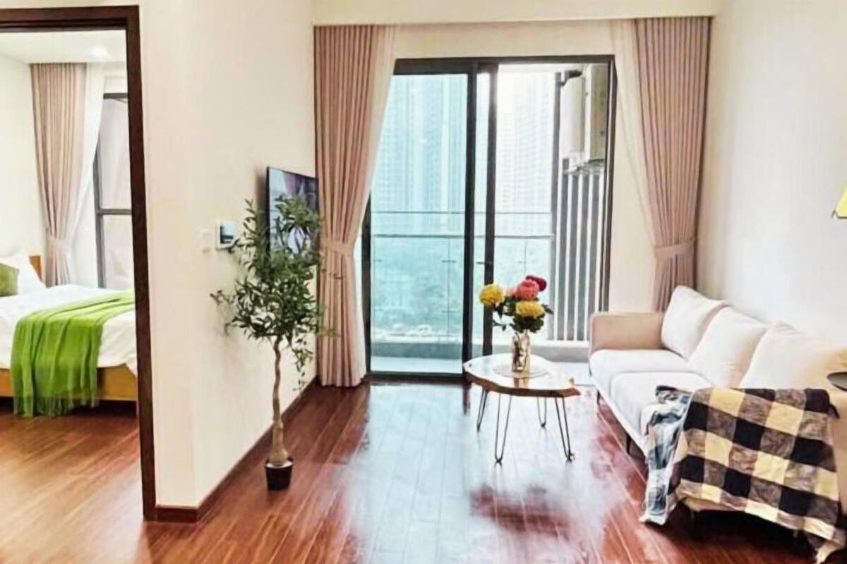 A hotel room with a wooden floor, a sofa, a small table with a vase, and a view through a large window.