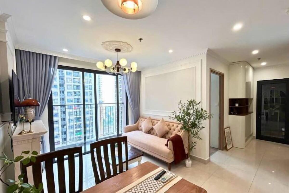 A modern living room with a sofa, dining table, and chairs, a large window with curtains, and a chandelier.