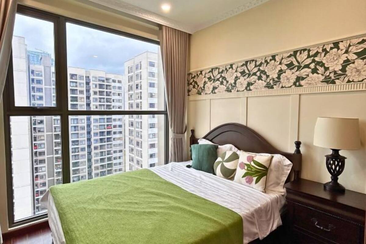 A hotel room with a large window, a bed with a green blanket, floral wallpaper, and a nightstand with a lamp.