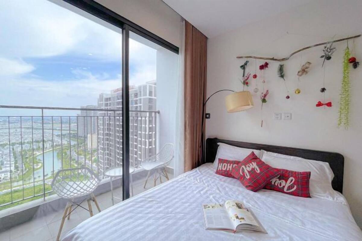 A hotel room with a large window, a bed with white bedding, a red and black pillow, a small table, and a chair.