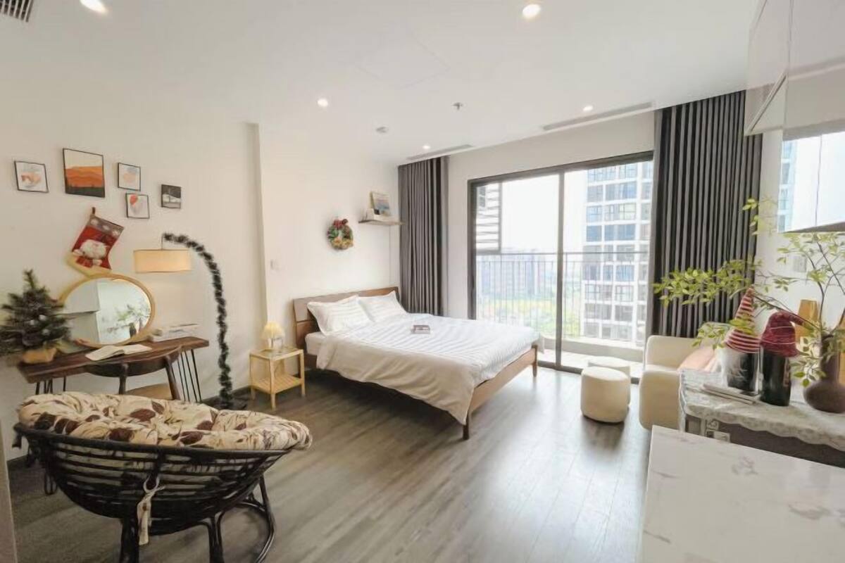 A modern hotel room with a bed, a desk, a chair, a small table, a plant, and a balcony with a view.