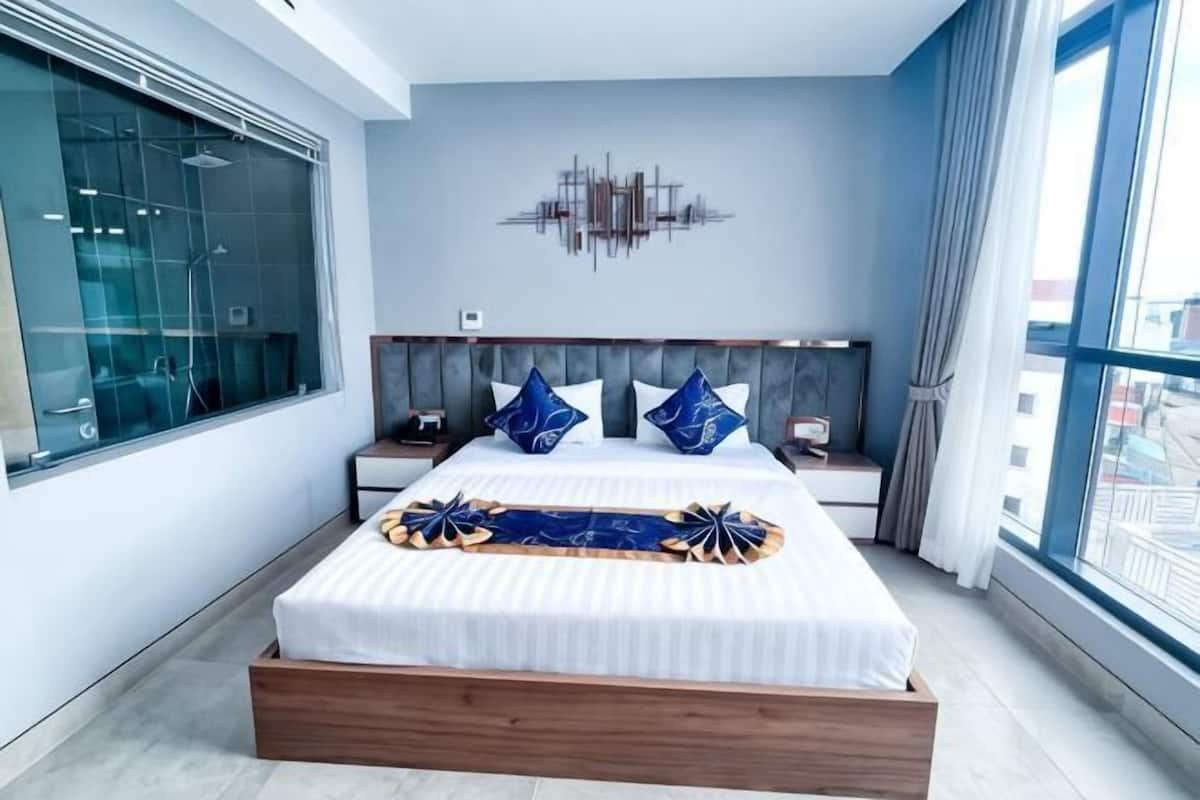 A modern bedroom with a large bed, a headboard with a light fixture, a nightstand, and a view of the city through large windows.