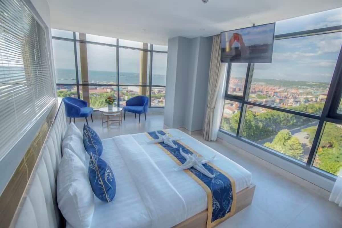 A hotel room with a large bed, two chairs, a small table, and a TV. The room has large windows offering a city view.