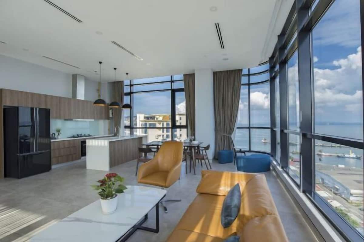 A modern living space with a kitchen, dining area, and seating by large windows overlooking a cityscape.