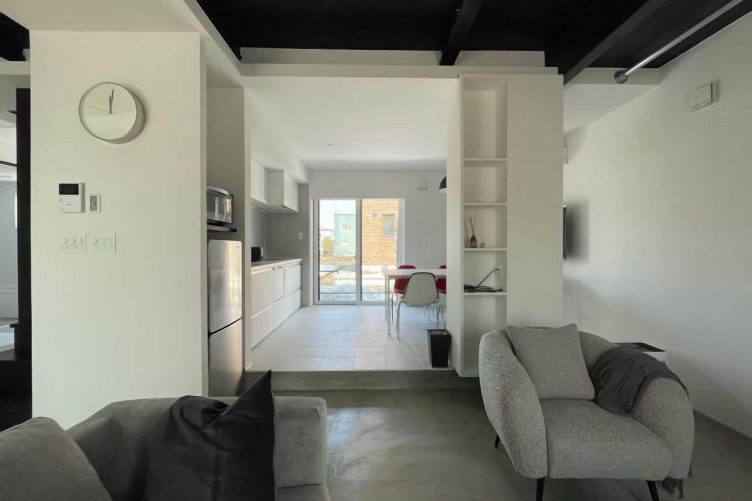 A modern living room with a sofa, armchair, and a flat-screen TV. A kitchen area with appliances is visible in the background.