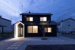 A two-story residential building with a dark exterior, a visible entrance, and a small tree in the front yard. - Biei Villa Gen (Kamikawa District)