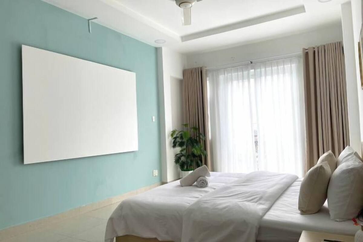 A hotel room with a bed, a large window with curtains, a ceiling fan, and a wall-mounted TV.