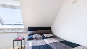 Een slaapkamer met een bed, een raam, een klein tafeltje en een stopcontact aan de muur.