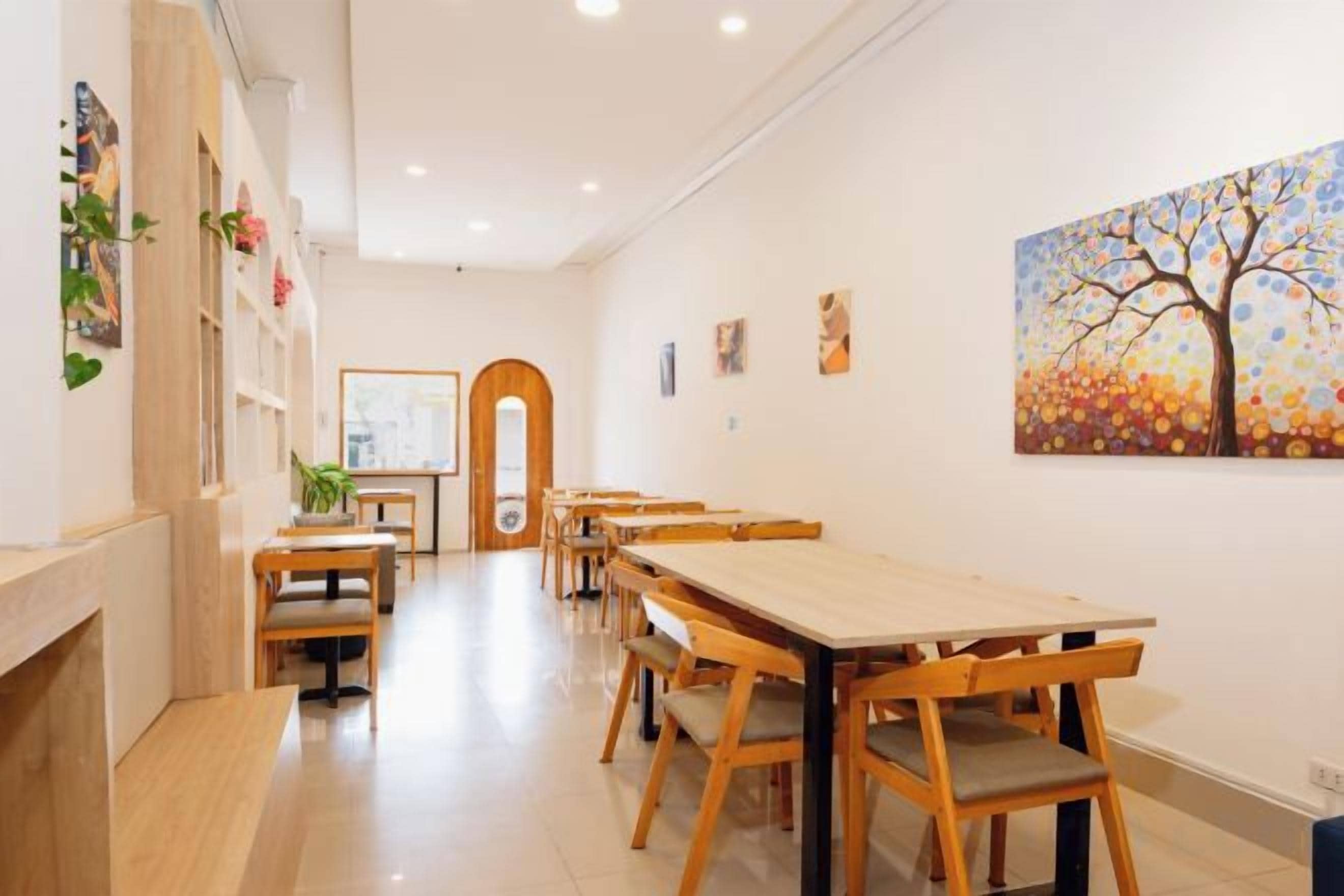 Un espace repas avec des tables et des chaises en bois, un mur blanc orné d’un tableau, et une porte en bois à l’arrière-plan.