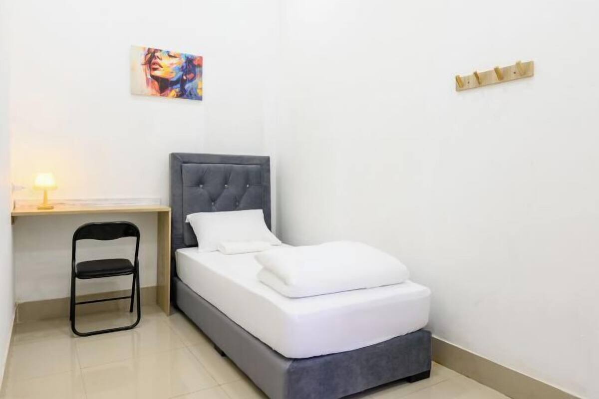 A small, neatly arranged bedroom with a bed, a chair, a desk, and a painting on the wall.