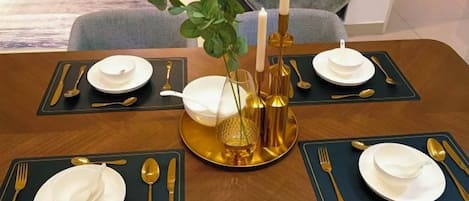 Mesa de comedor dispuesta para cuatro personas, con individuales dorados y negros, platos blancos y cubiertos dorados.