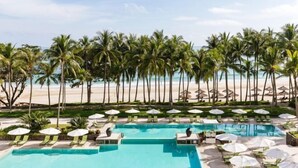 A pool area with multiple swimming pools, lounge chairs, and umbrellas, surrounded by palm trees and a beach in the background. - Club Med Bintan (Bintan)