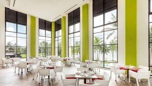 A spacious indoor dining area with multiple tables and chairs, large windows with green vertical accents, and a view of palm trees and a beach. - Club Med Bintan (Bintan)