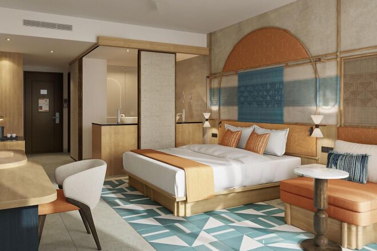 Una habitación de hotel moderna con una cama grande, un área de estar con un sofá y un sillón, una mesita y una pared decorativa con patrones geométricos.