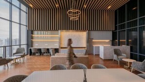 Een modern interieur met een hoge plafond, houten latten en een centrale lichtarmatuur. Er zijn zithoeken met tafels en stoelen, en een groot raam met uitzicht op de stad.