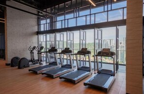 Une salle de sport équipée de tapis de course, de vélos elliptiques et de vélos d’appartement, avec de grandes fenêtres offrant une vue sur la ville.