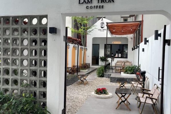A café entrance with a sign reading 'LAM TRON COFFEE,' outdoor seating with tables and chairs, and a small garden area with flowers.