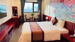 Ein Hotelzimmer mit einem großen Bett, einem Schreibtisch mit Stuhl, einem Fernseher, einem Bücherregal und einem fenster mit Vorhängen.