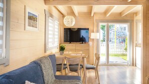 A modern living room with a wooden floor, a blue sofa, a dining table with chairs, a TV, and a large window with a view of a playground. - Collins Resorts are dream vacations for families with children right on the beach. (Sianożęty)