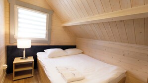 A wooden room with a bed, a nightstand, a lamp, and a window with blinds. - Collins Resorts are dream vacations for families with children right on the beach. (Sianożęty)