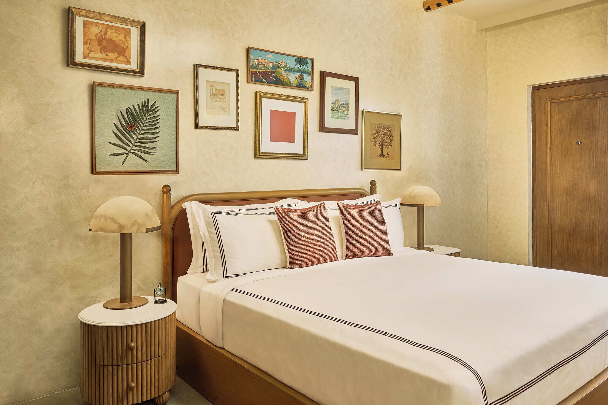 Una cama bien tendida con un cabecero de madera, flanqueada por dos mesitas de noche con lámparas, en una habitación con pared beige texturizada y cuadros enmarcados.