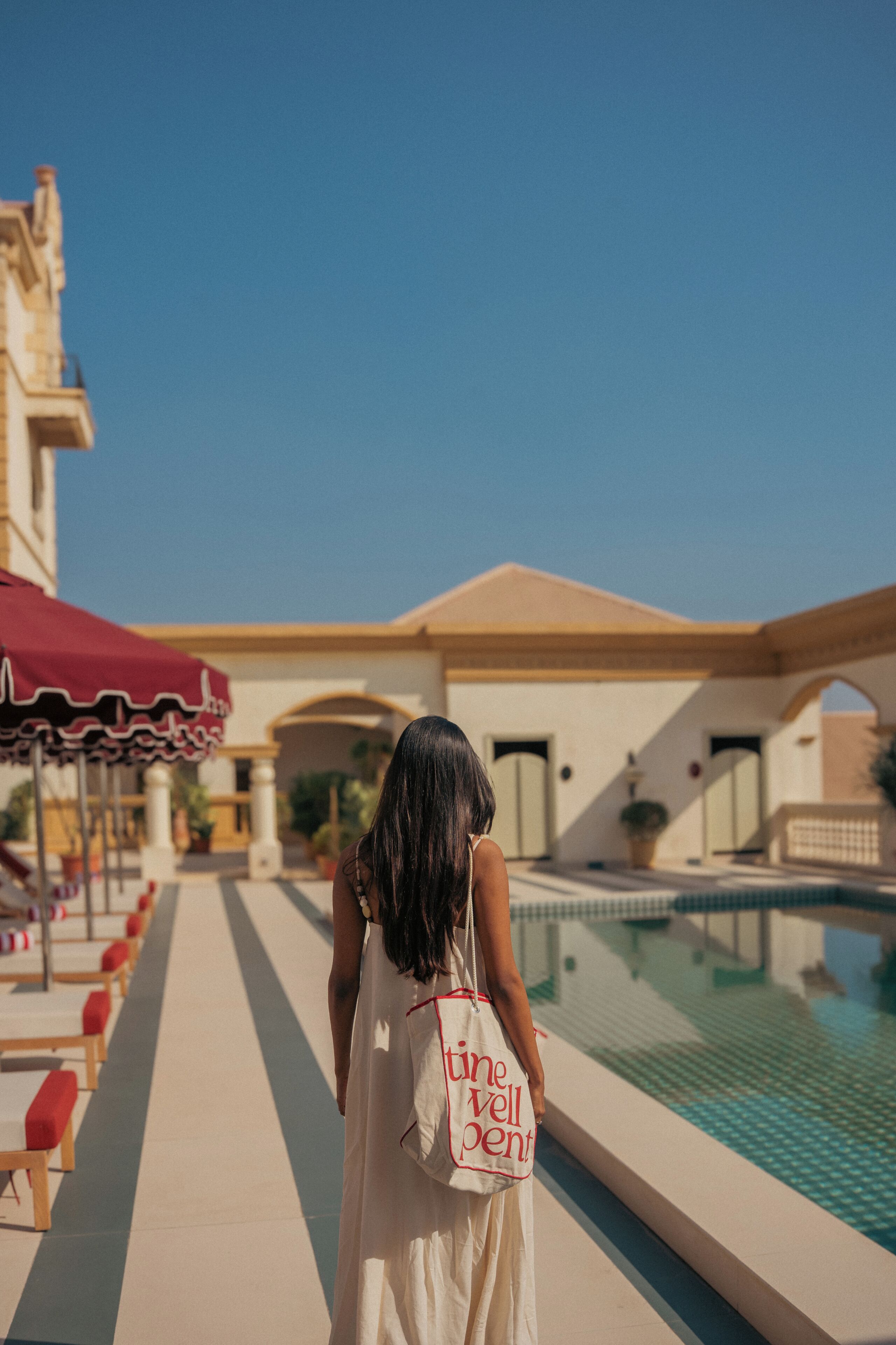 A woman in a white dress walking along a poolside pathway with red benches and a red umbrella.