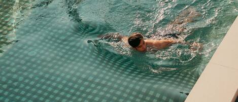 A pool with a checkered pattern and a person swimming. There is a towel and a drink on the poolside.