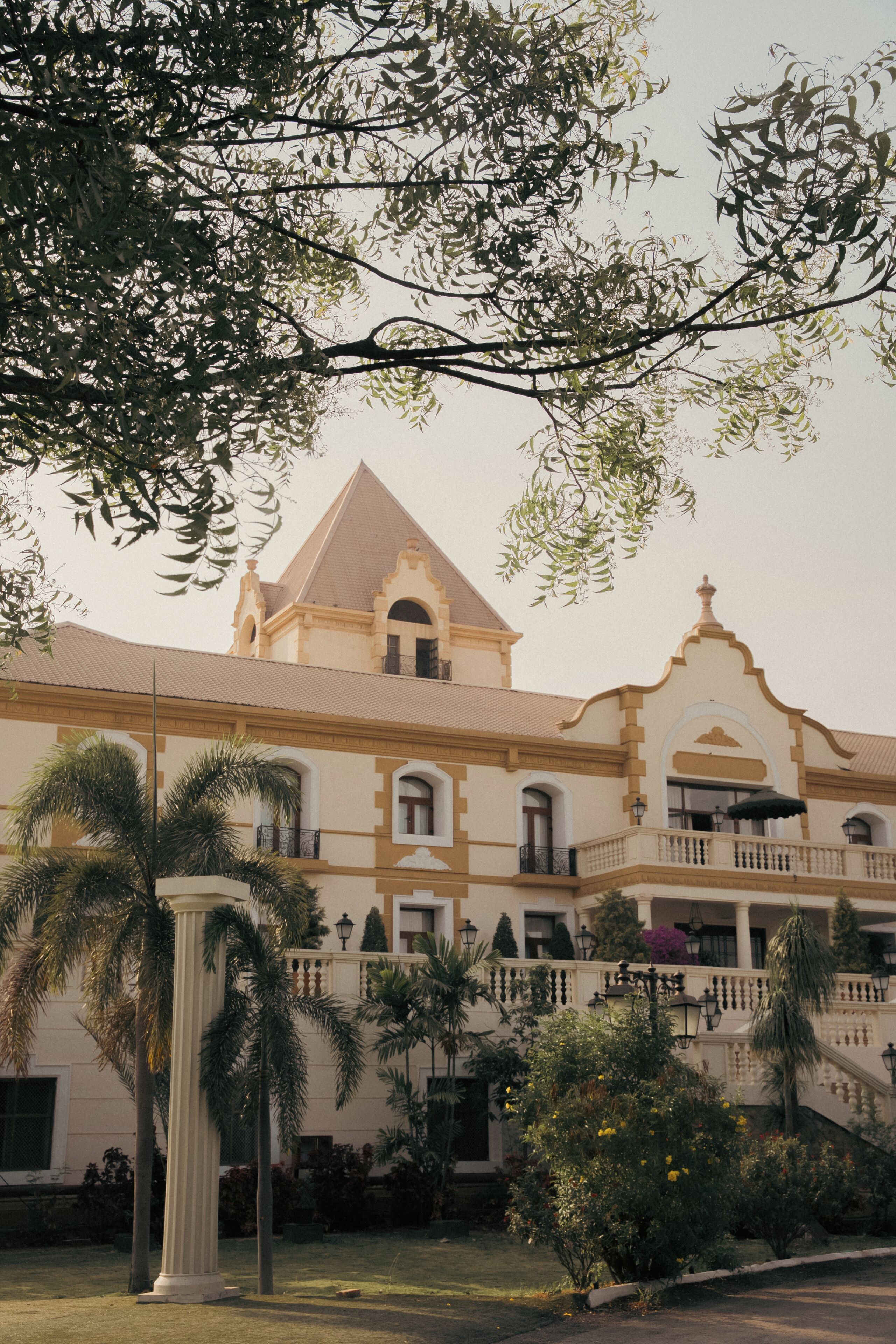 A large building with a prominent central tower, multiple balconies, and a surrounding garden with palm trees.