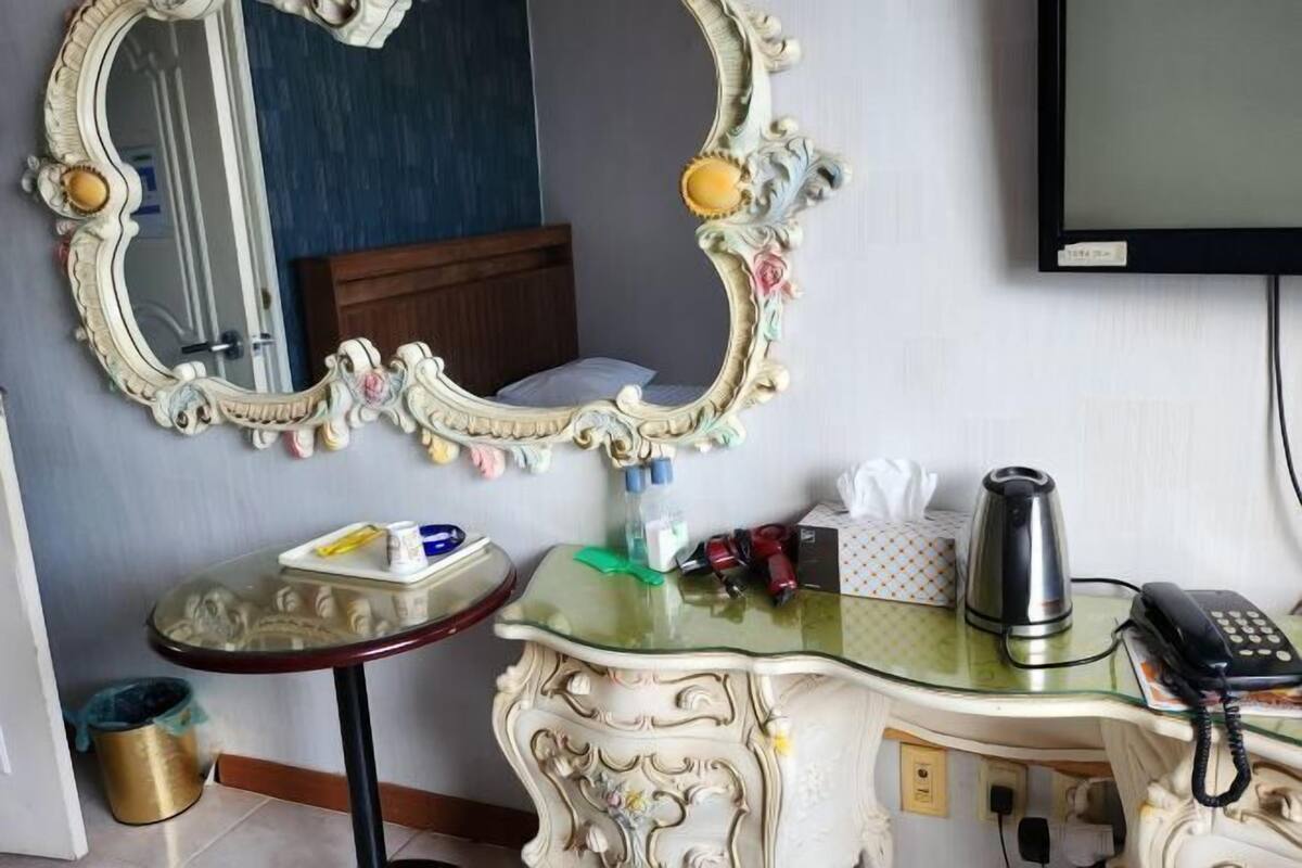 A room with a large ornate mirror, a small round table with a glass top, a bedside table with a telephone, and a television mounted on the wall.