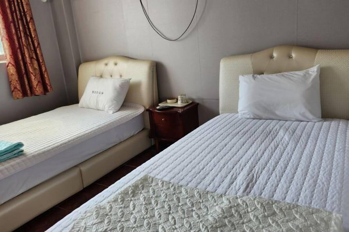 A hotel room with two beds, a bedside table, a wall-mounted light, and a curtain.