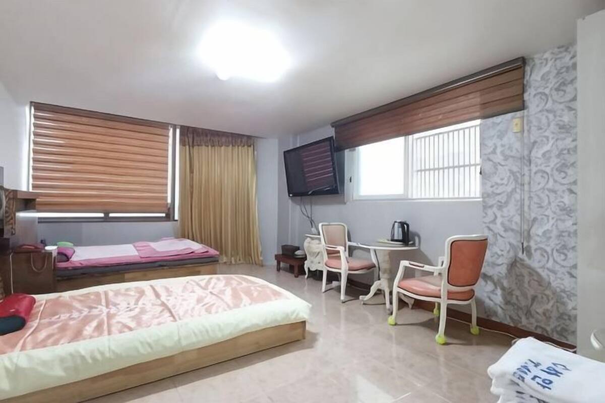 A room with a bed, a desk with chairs, a television, and a window with blinds.
