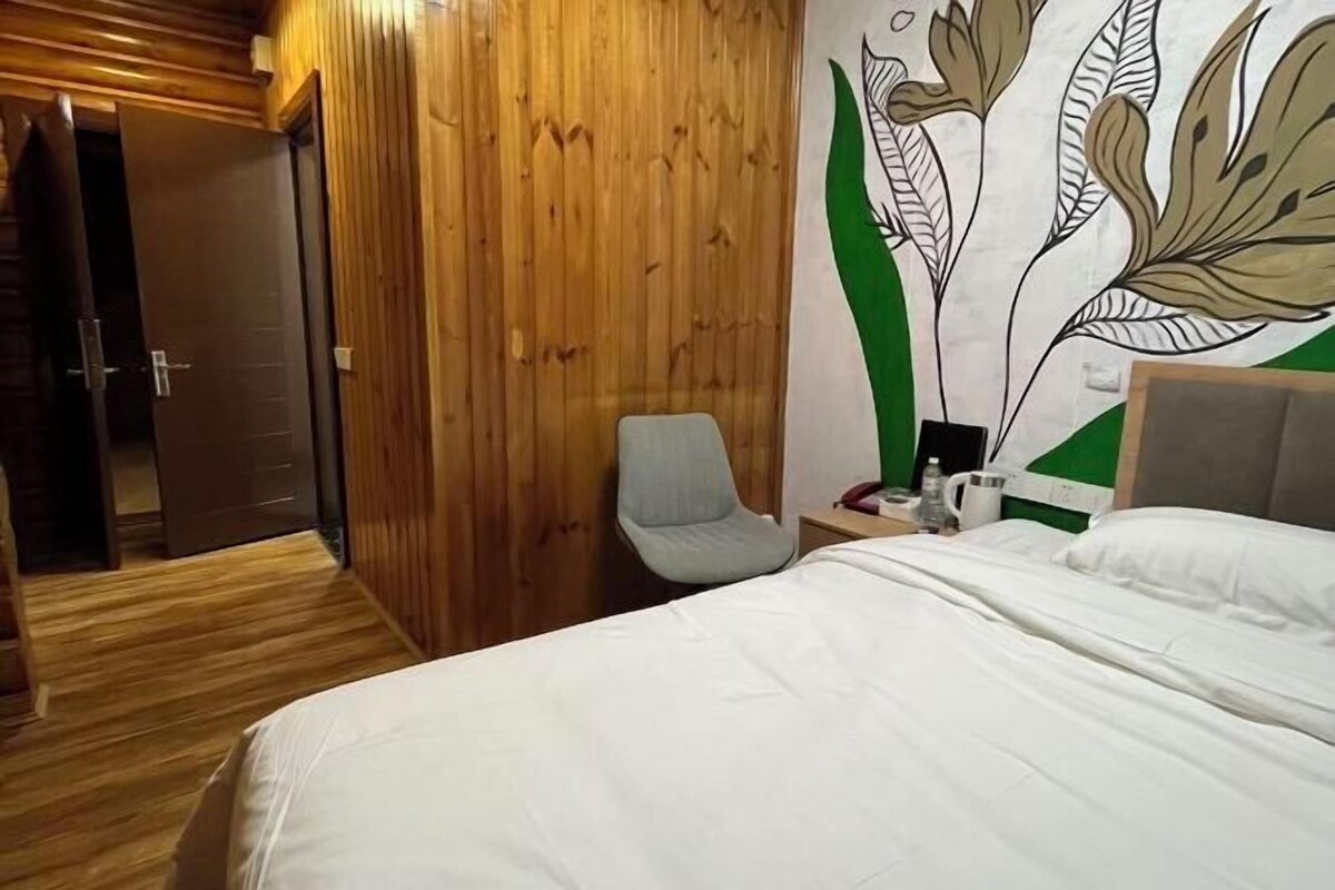 A hotel room with a wooden wardrobe, a bed with white bedding, a grey armchair, and a wall with a floral mural.