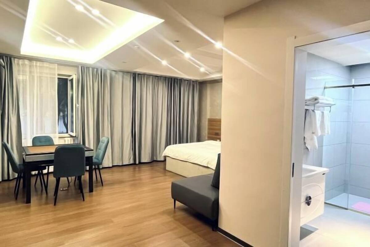 A hotel room with a wooden floor, a bed, a desk with chairs, a sofa, a shower area, and a large window with curtains.