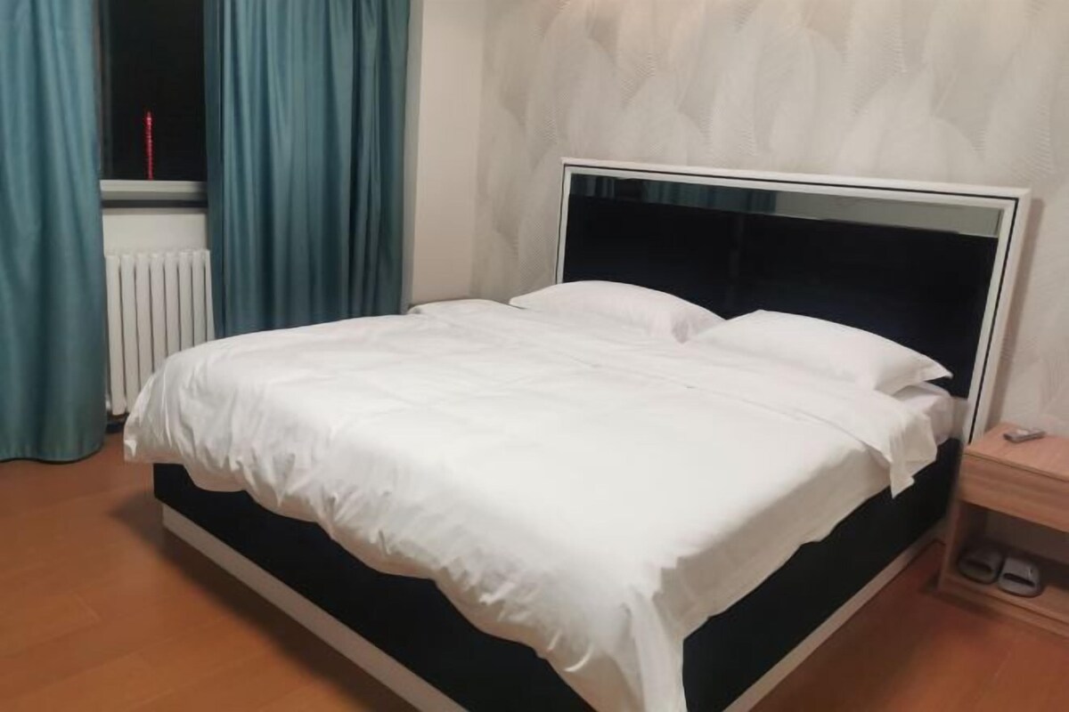 A bed with white bedding, a wooden nightstand with a remote, and a window with curtains.