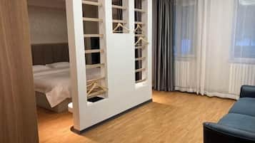 A bedroom with a bed, a wardrobe with hangers, a window with curtains, and a wooden floor.