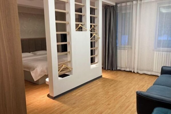 A bedroom with a bed, a wardrobe with hangers, a window with curtains, and a wooden floor. - Yingbin Garden Hotel (Almaty)