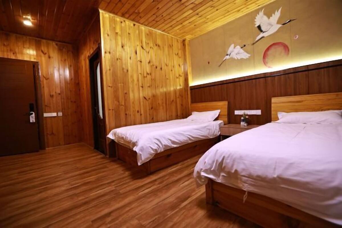 A room with wooden floors and walls, two beds with white bedding, a wooden door, and a wall with a bird and moon mural.
