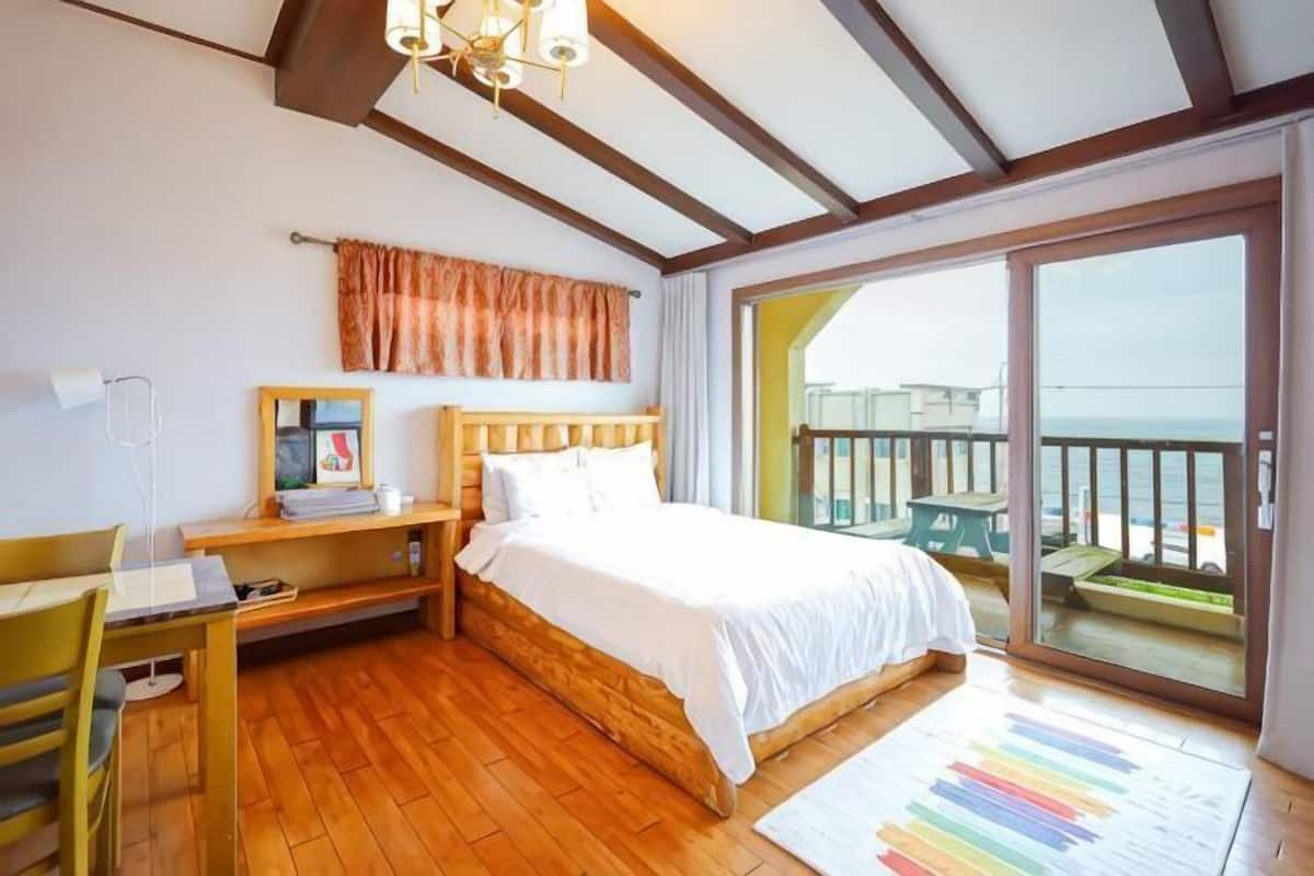Un dormitorio con cama de madera, un escritorio, una silla, un ventanal y vista al mar.