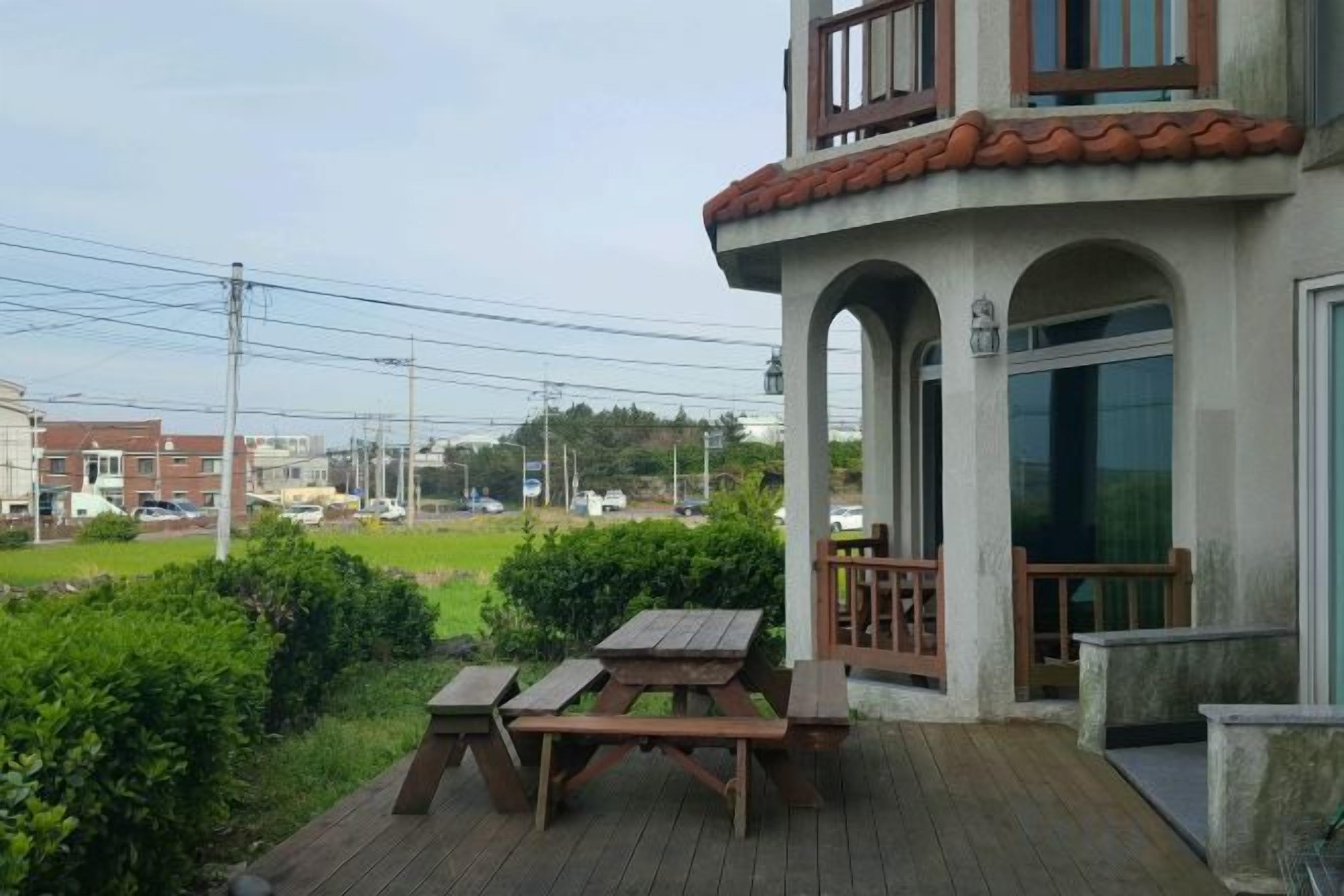 A building with a covered outdoor area featuring a wooden picnic table and benches, a balcony with a railing, and a view of the surrounding area.