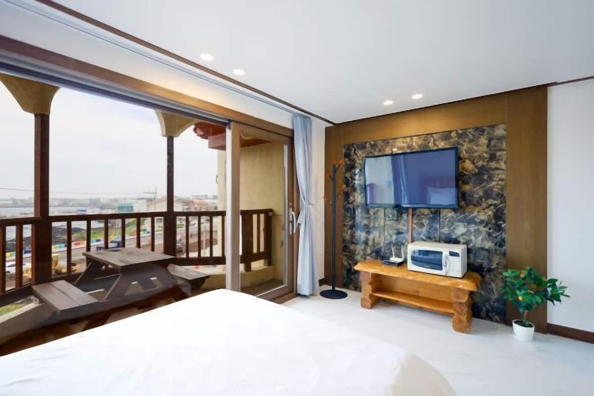 Habitación de hotel con cama, televisor montado en pared de piedra, una mesita con microondas, balcón con vistas a la ciudad y una planta en maceta.