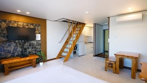 Un dormitorio con una cama, un banco de madera, una mesita, un televisor montado en una pared de piedra y una escalera que lleva a una cocina.