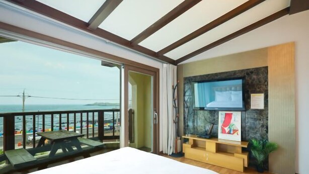 Un dormitorio con una cama, un televisor montado en una pared de piedra, un banco de madera y un balcón con vistas al mar.