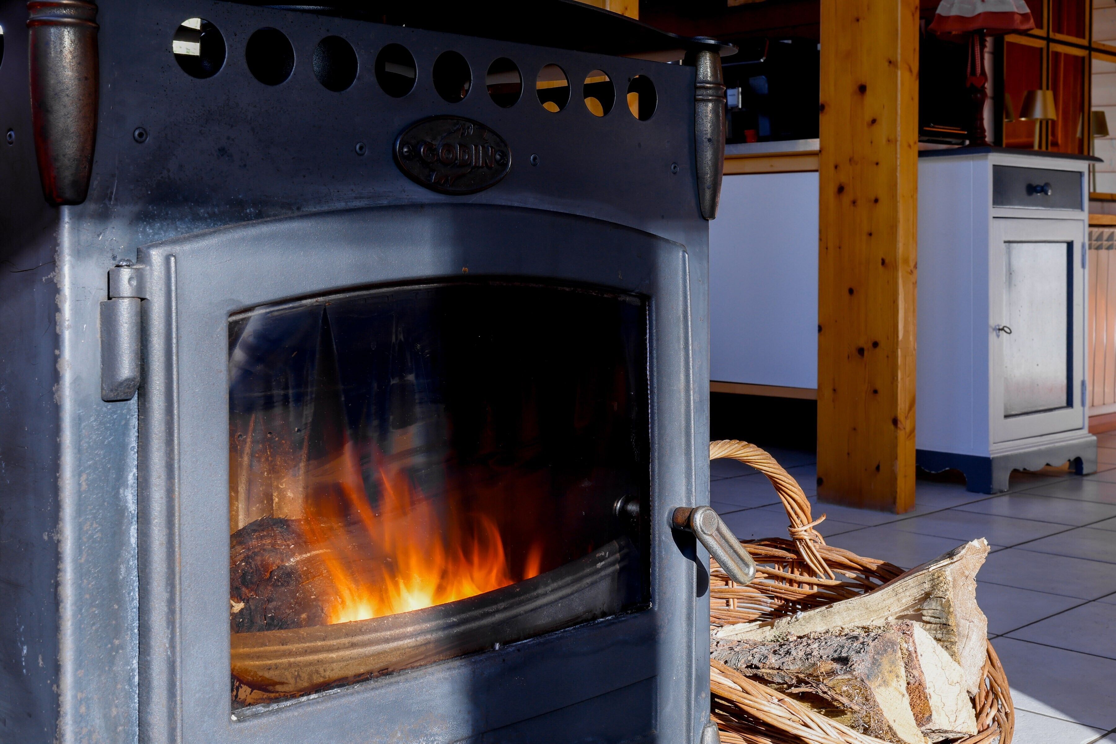 Lareira a lenha com porta de vidro, cesta de vime com lenha e área de cozinha com armários.