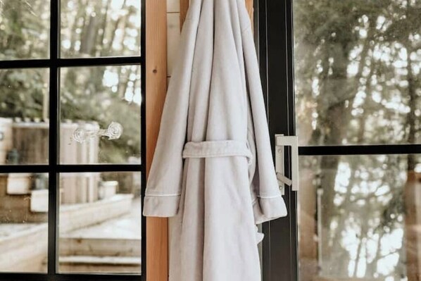 A window with a view of a wooden deck and trees, and a white robe hanging on a rack.